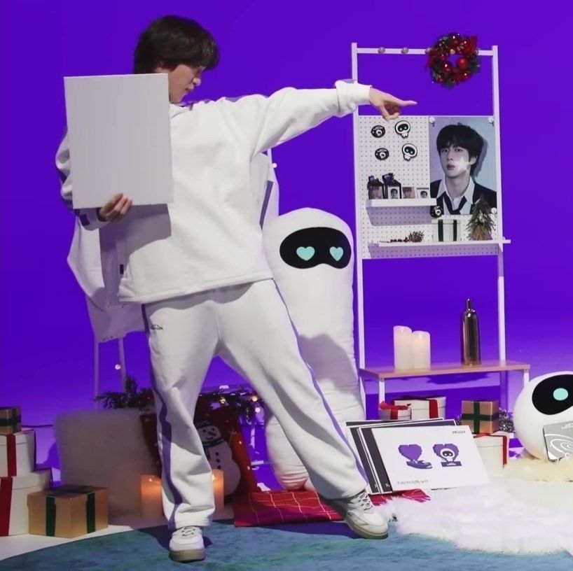 Jin of BTS pointing his index finger towards JHope he's wearing a white pantsuit with purple stripes on the sides on the set of wootteo merch video