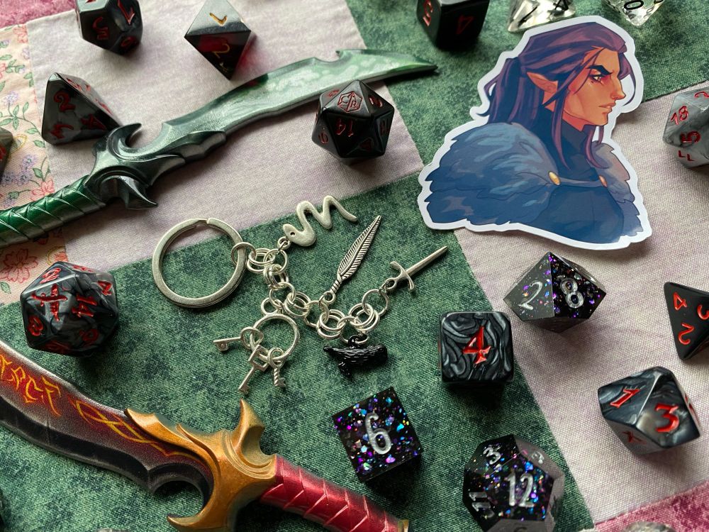 A keychain on a quilt surrounded by polyhedral dice, a sticker of vax from critical role, and two daggers. The keychain features charms of a dagger, a crow, a feather, a keyring, and a snake.