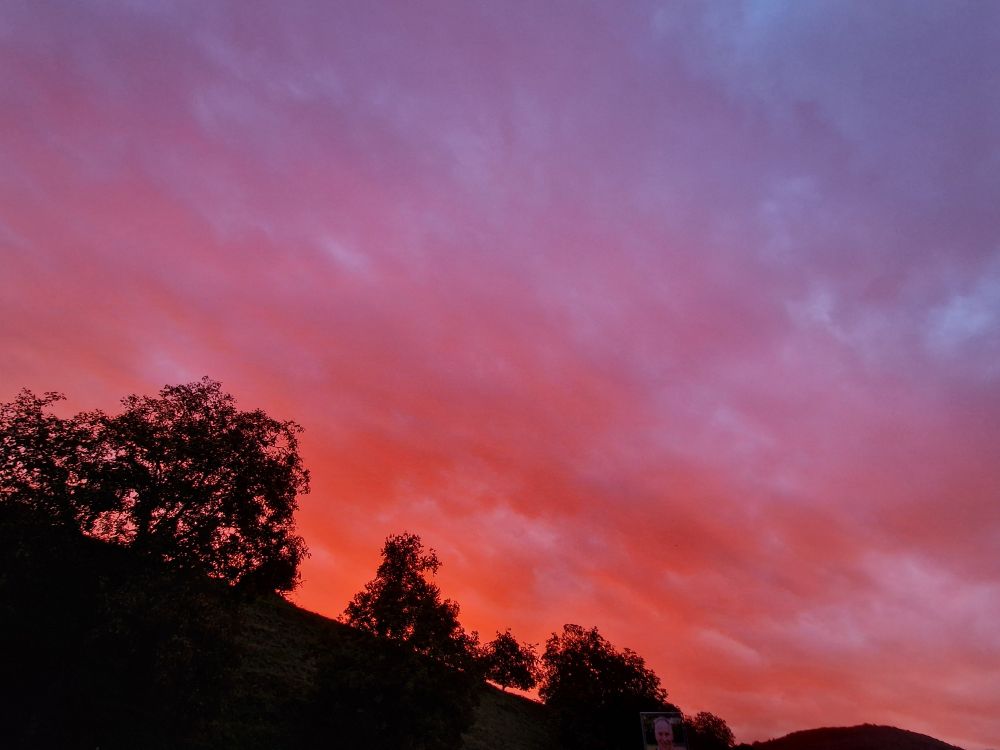Dieses Foto zeigt einen dramatischen Sonnenaufgang über einem Hügel in einer ländlichen Gegend. Der Himmel ist in leuchtendes Rot und Magenta getaucht, eine Farbpalette, die den größten Teil des Bildes ausfüllt. Weiter oben am Himmel gehen die Farben in einen kühleren Blau- und Lilaton über. Der Horizont ist durch einen bewaldeten Hügel definiert, dessen Silhouetten klar gegen den leuchtenden Himmel zu sehen sind. Die Bäume sind dunkel und scheinen von den warmen Farben des Sonnenuntergangs angestrahlt zu werden. Ein beleuchtetes Plakat mit einem Porträt, wahrscheinlich ein Wahlplakat, ist am unteren rechten Rand des Bildes zu sehen und fügt einen menschlichen Berührungspunkt in diese ansonsten naturbetonte Szene ein. 