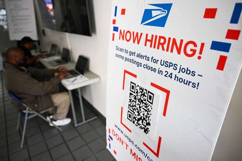 A now hiring sign is displayed as job seekers fill out applications for employment at a United States Postal Service (USPS) job fair hiring automotive technicians and motor vehicle operators at the Los Angeles Processing and Distribution Center in Los Angeles, California on May 16, 2025. (Photo by Patrick T. Fallon / AFP) 