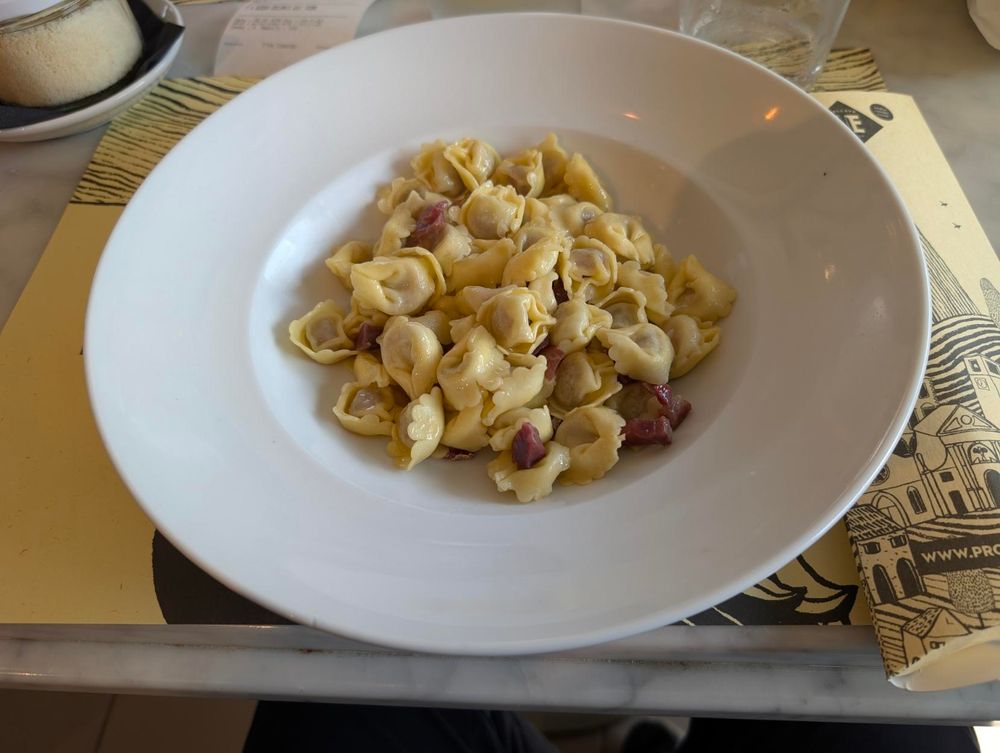 A high-angle shot shows a white bowl filled with tortellini, some of which have visible bits of prosciutto San Daniele. The bowl is centered on a placemat with a black and white drawing. A small white bowl with parmigiano is partially visible in the upper left corner.