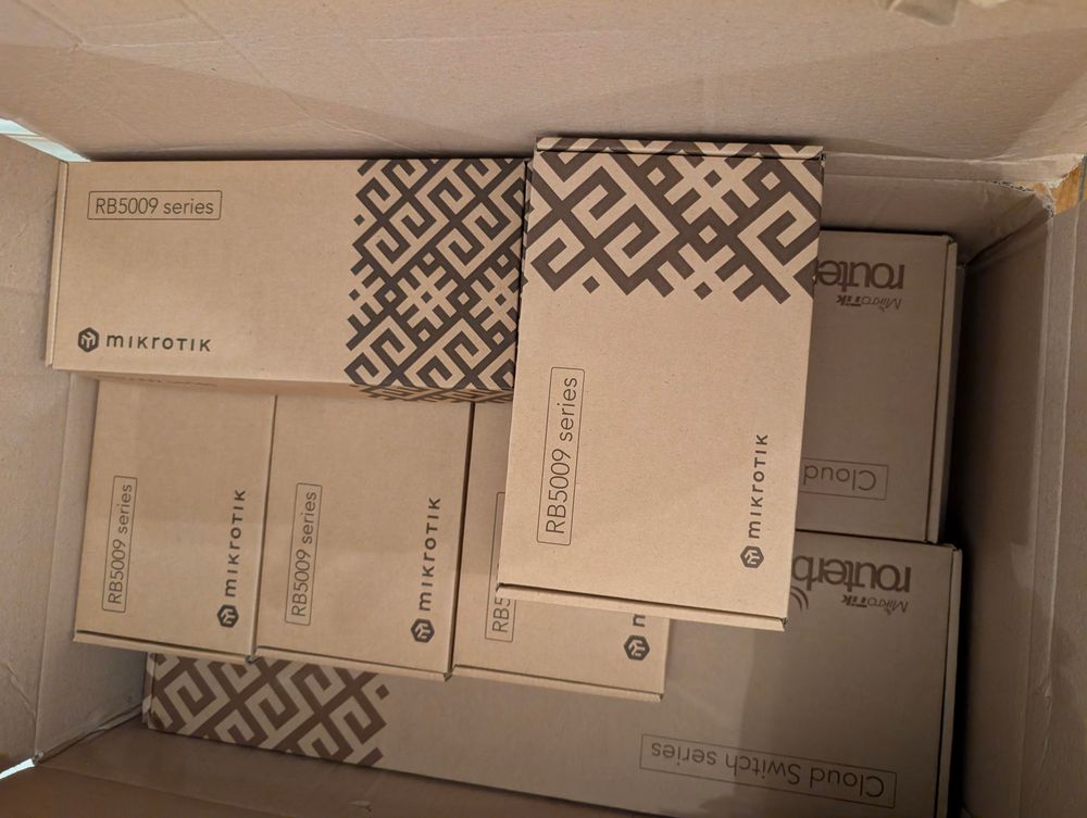A large cardboard shipping box filled with multiple Mikrotik product packages. Most of the boxes are labeled “RB5009 series” with the Mikrotik logo and a geometric patterned design. Additional Mikrotik boxes, including some from the “Cloud Router Switch series”, are partially visible underneath. The boxes are neatly arranged and are new and unopened.
