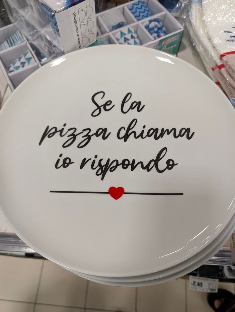 A close-up of a white decorative plate with the Italian text "Se la pizza chiama io rispondo" in black cursive font, which translates to "If pizza calls, I answer." A small red heart is positioned on a thin black line below the text. The plate is stacked on other identical plates, and in the background, other household items are visible on a store shelf.