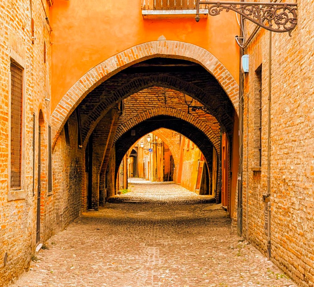 A long, narrow, cobblestone alleyway in Via Delle Volte, Ferrara, Italy is lined with ancient brick buildings. A series of deep brick arches creates a tunnel-like effect, leading the eye down the path. The warm, rustic tones of the old bricks and the golden light evoke a sense of timelessness and history.