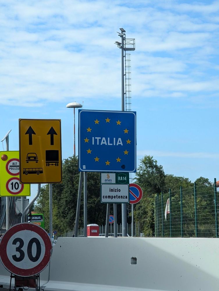 A full outdoor shot of a collection of street signs. The most prominent sign is a square blue sign with the word "ITALIA" and the European Union's circle of yellow stars. Below it is a smaller white sign that reads "Inizio competenza" and another with "RA 14" in green text. To the left is a large yellow sign with two black arrows pointing upwards, indicating separate lanes for cars and trucks.