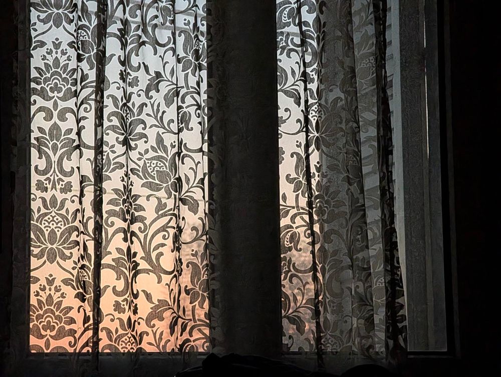 Semi transparent patterned curtains hang in front of a window before sunrise, with soft early light glowing through the fabric and revealing floral motifs while the room remains mostly dark.