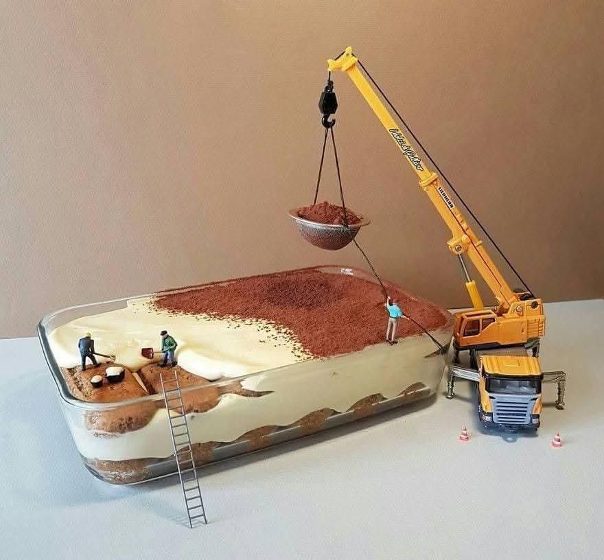 A whimsical photo shows miniature construction workers "building" a tiramisu dessert. A toy crane lifts a container of cocoa powder over a large glass dish of tiramisu, while tiny figures on the dessert itself appear to be spreading cocoa and working on a section of ladyfingers with a ladder.