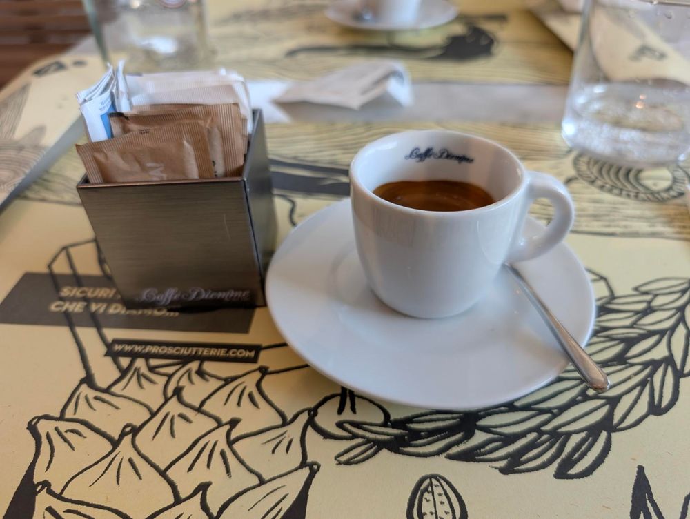 A top-down shot shows a small white cup of espresso with a spoon resting on its saucer, next to a silver sugar packet holder filled with various sugar and sweetener packets. They are sitting on a light-colored table with black drawings. A glass of water is visible in the background.