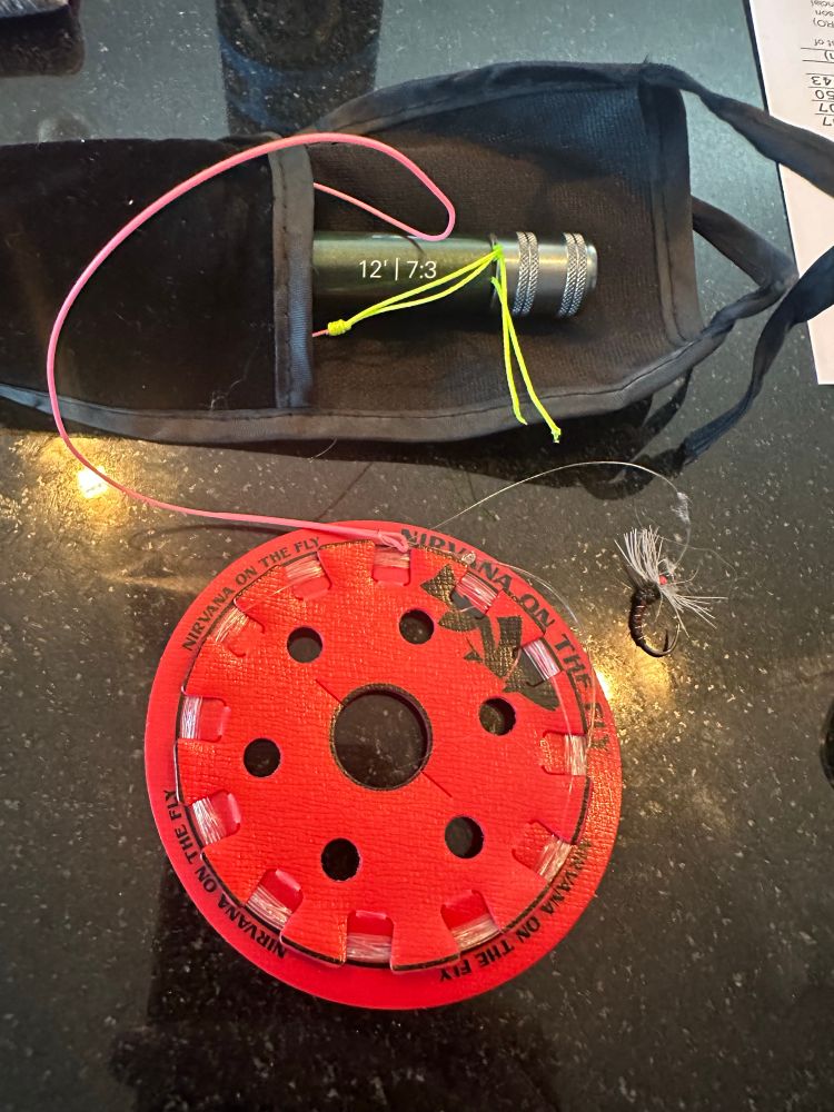 The tip of a tenkara rod poking out of its storage sleeve and a red tippet disc and fly.