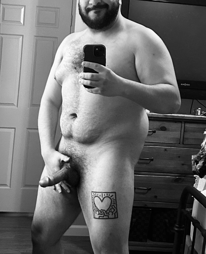 A B&W photo of Drew naked, his semi-hard cock and full bush on display as he holds his crotch and smiles for the camera