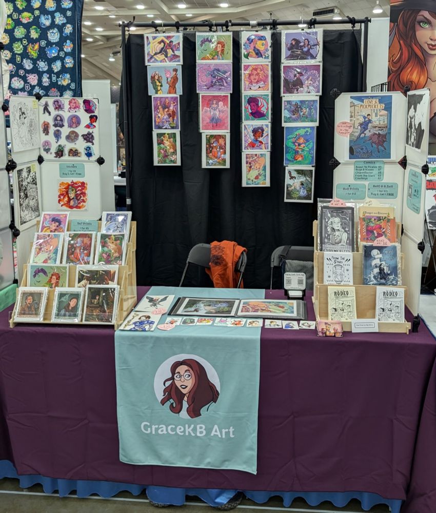 Booth setup with prints, stickers, comics, and sketches covering the table & large prints hanging behind