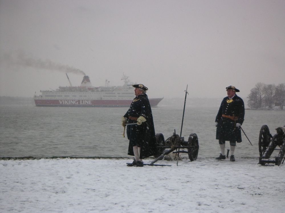 Two men in 18th century uniforms standing on a snowy shore with two cannons. In the distance there's a Viking Line cruise ferry going by.