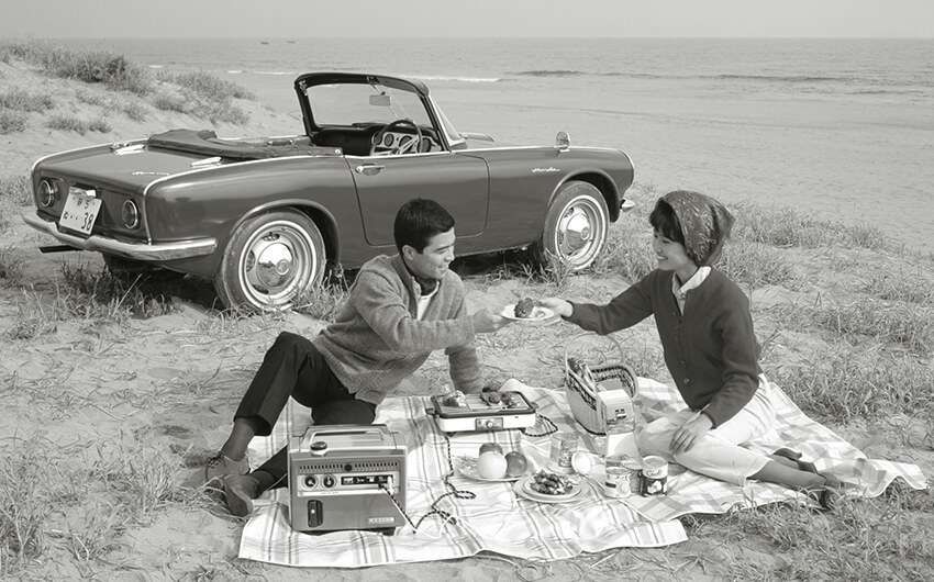 1960s black and white promotional photo for the Honda E300 portable generator showing a picnicking couple using one at the edge of a beach with a Honda S600 car behind them
