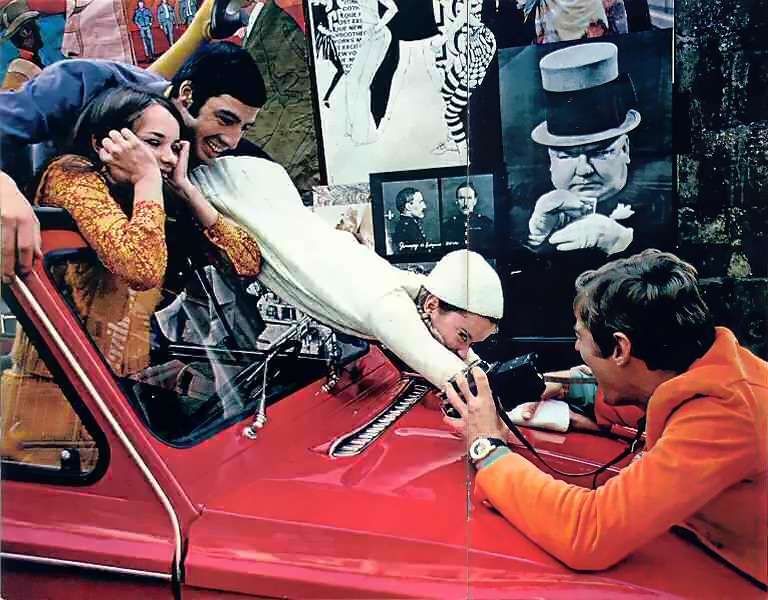photo from an old brochure of some groovy young people with a Citroen Dyane you can't see much of because it's in closeup, three of the people are stood up in the front out of the sunroof and one of those ls sliding forward onto the bonnet, leaning across the bonnet from the other side is a guy holding a camera like he's David Hemmings in some badly dated groovy film, on the wall behind them are groovy pictures of Edwardians and shit