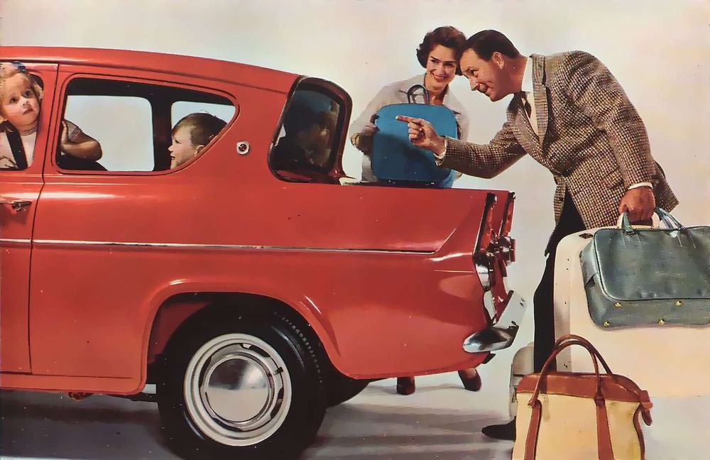 1964 brochure photo of  a side view of the rear of a red Ford Anglia in a white studio, there's kids in the car and a man and woman behind it with suitcases, she's smiling at them and he's pointing at them for no apparent reason, none of the kids appear to be looking at them