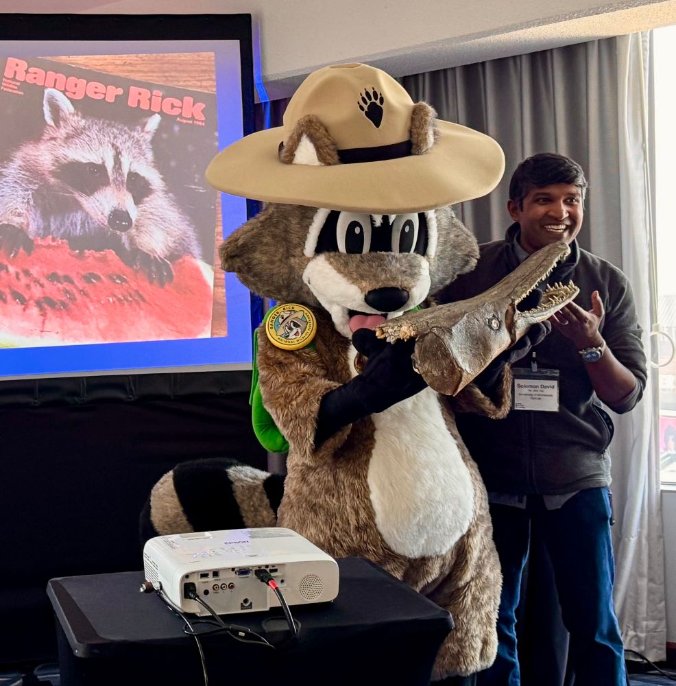 A fish scientist stands next to Ranger Rick, the raccoon mascot of the nature magazine that inspired him as a kid and introduced him to the fish (gar) that he’d make his career studying and trying to conserve (as a broader effort for freshwater conservation). They are holding up an Alligator Gar head, and the scientist still can’t believe this happened!!!