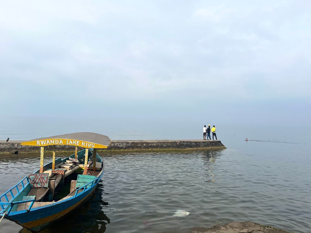 Three men stand at the end of a pier looking out to the lake. In the foreground is a small blue tourist boat with a partial yellow roof with the words Rwanda Lake Kivu. 