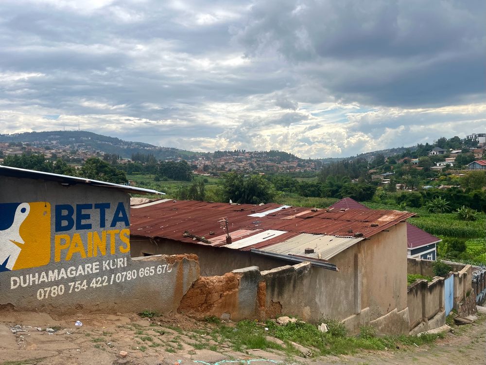Rows of informal houses with tin roofs descends down the hall. The first has a blue and yellow sign saying ‘Beta Paints’. In the distance, multiple hills give way to a dramatic cloudy sky