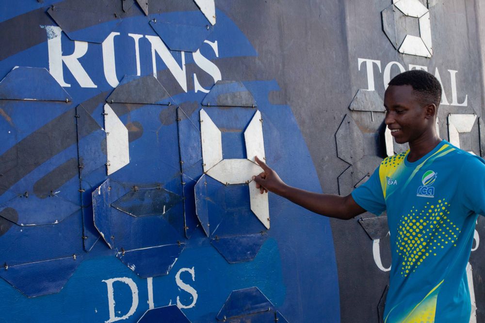 A young Rwandan man in a blue and yellow patterned t-shirt, turns over the leaves of the blue manual scoreboard which shows the words ‘Runs’ and ‘Total’. His forefinger rests on the white number 4, as he looks down and away from the camera with a smile. 