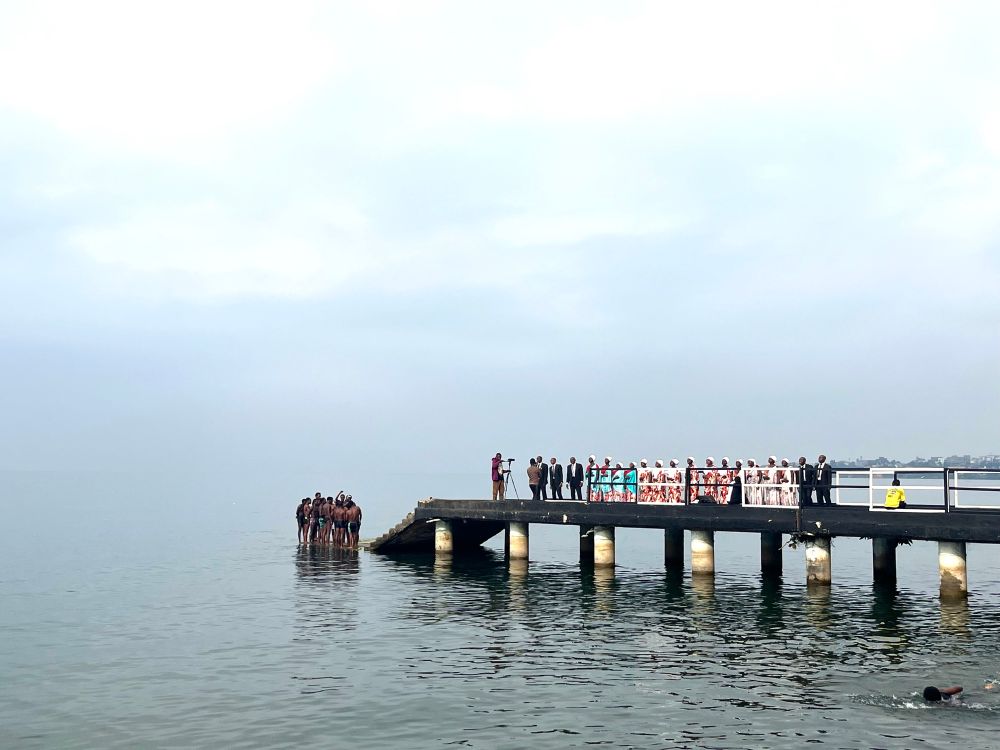 A wedding party in matching outfits is filmed performing a song for the camera, on the pier on lake Kivu, Gisenyi. A group of male swimmers huddle on the neighbouring platform nearby. 
