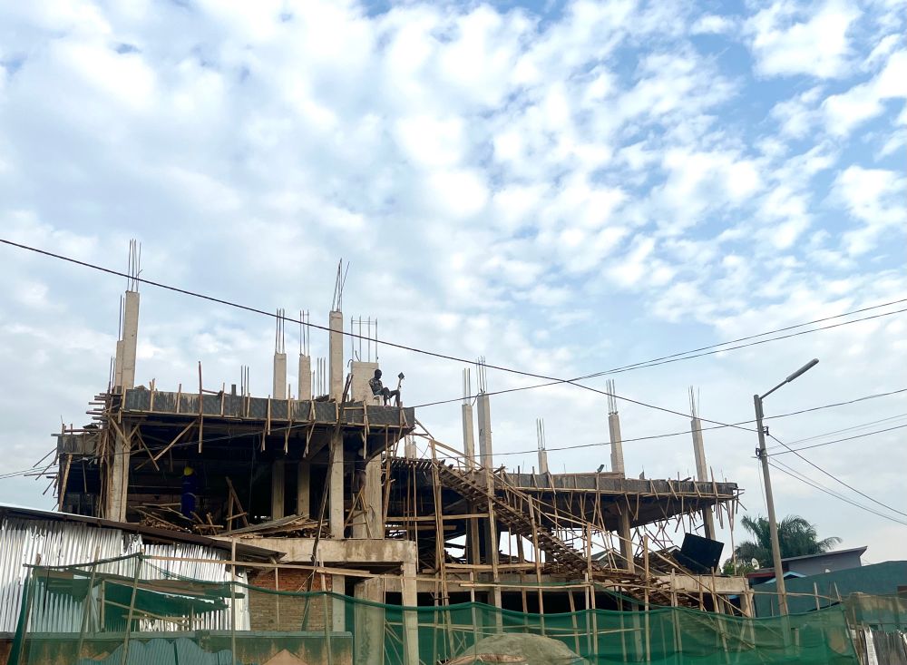 A building site of two storeys, with no walls, just upright concrete pillars and floors. A young man sits on a chair on the corner of the second storey. The sky behind is blue with white fluffy clouds. There is a green mesh fabric fence in front of the building site. 