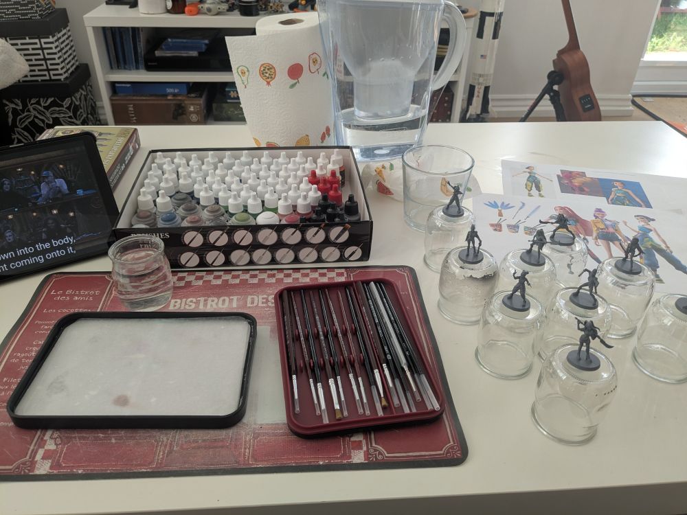 My whole painting set-up, with brushes, paints, a wet palette, and all the minis fixed on top of glass pots. There's also a tablet with critical role on it, and some papers with the Huntrix girls printed on it for color reference.