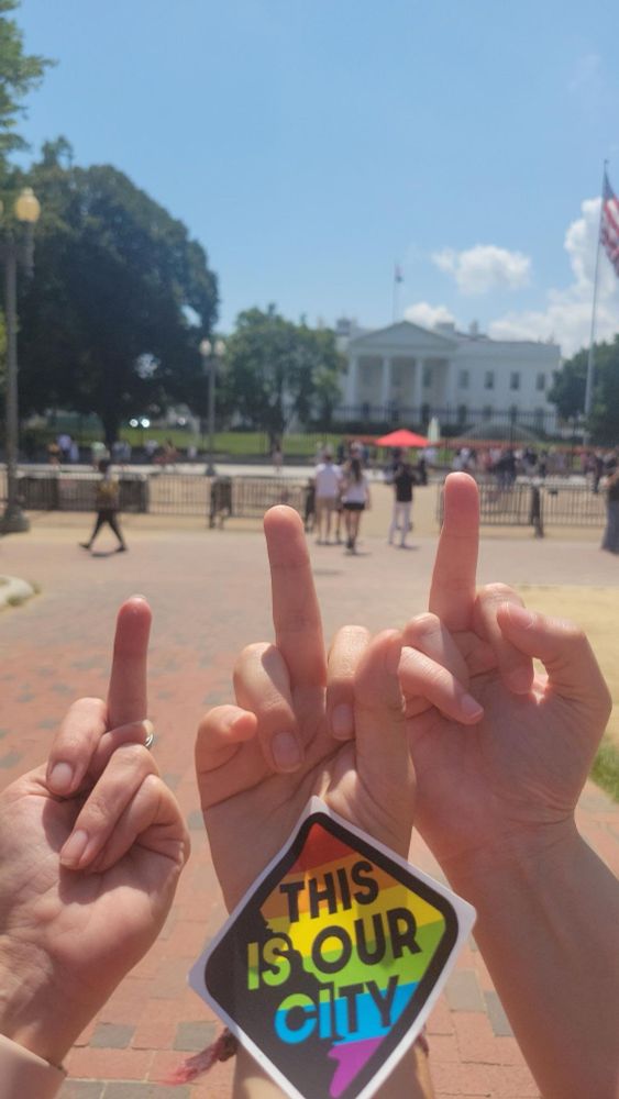 3 hands giving the middle finger (ie, saying “F*** you”) to the White House, with a sticker that says “THIS IS OUR CITY” over a rainbow DC.