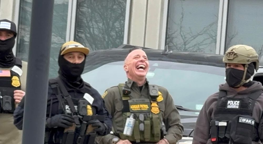 Greg Bovino laughing in front of protesters, standing next to multiple ICE agents in masks.