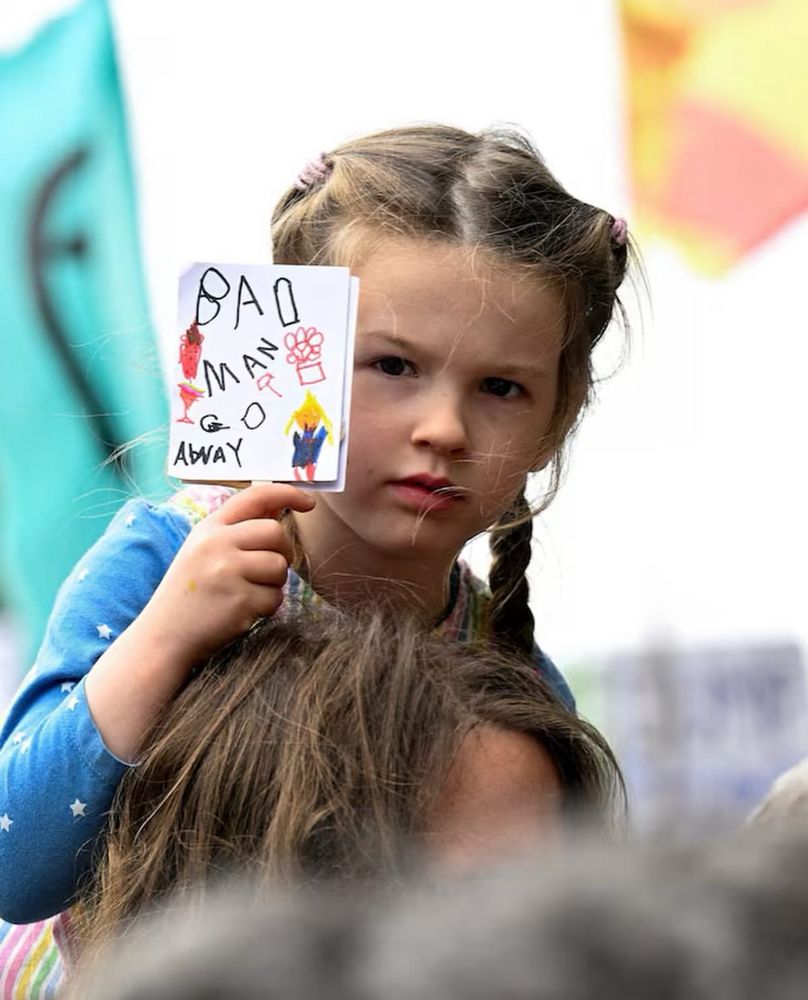 Photo of a young Scottish girl riding on the shoulders of her parent holding up a card that looks like a child drew it, which says “bad man go away.”