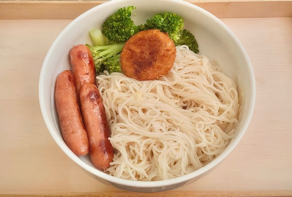 木のお盆の上に乗せた白い丼の中に水気を切り麺つゆをかけた素麺が入っている。
素麺の上には12時の方向に茹でたブロッコリーと小さなコロッケ。
9時の方向にソーセージが乗せてある。