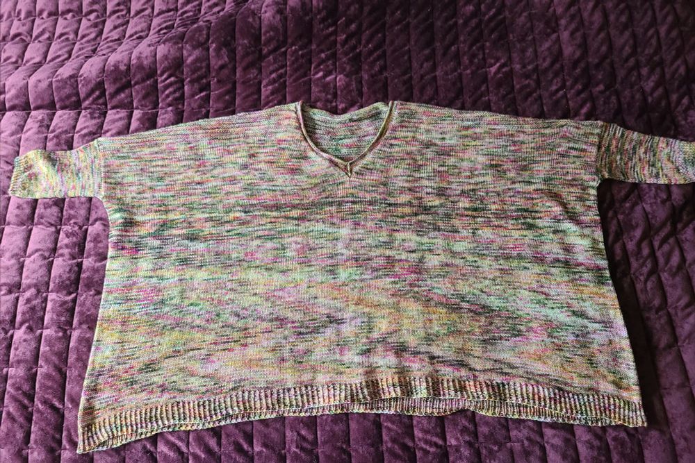 A v-neck wide jumper with very short, small sleeves laying on a maroon background. The jumper is made of variegated yarn in greens, pinks, yellows, and orangey bits.