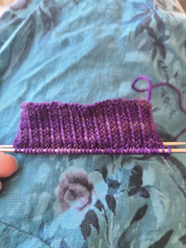 A 2 x 1 rib in deep purple sock yarn, the shortest of cuffs for a pair of socks. The background is a deep teal linen covered in a floral pattern of lighter and darker teals.