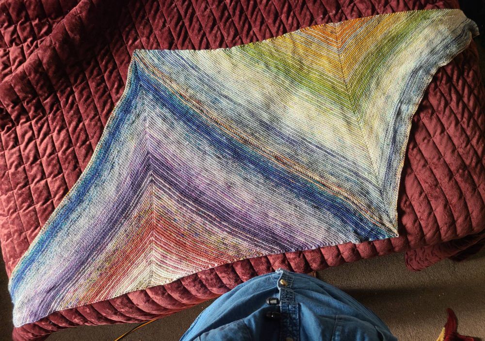 A knitted shawl made from two triangles joined together at the sides to create a parallelogram. Coloured vary from speckled creams, yellow, pink, blues in a rainbowy sequence almost.