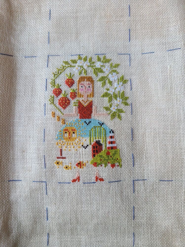 A cross stitch of a woman holding a white flowering bough over her head with strawberries hanging off. The skirt of her dress forms an idyllic summer beach scene with a boat, fish, and a lighthouse on a hill. The sun shines down on everything.