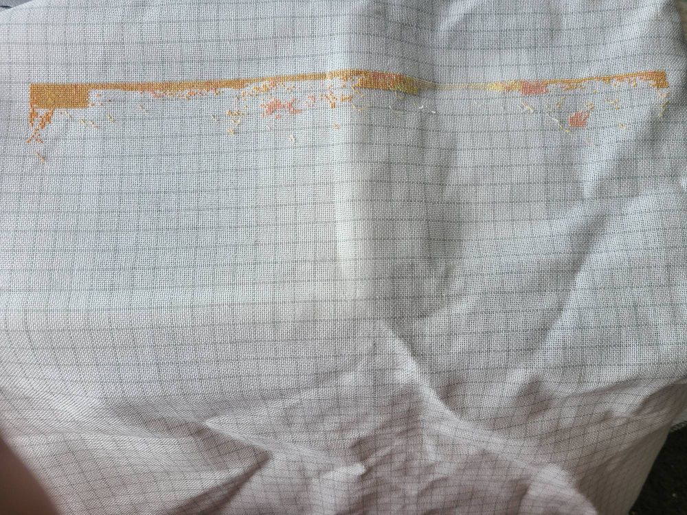 White evenweave cross stitch fabric covered with a gray 10 x 10 grid and some stitches across the top in tans, pinks, and yellows that will become the sky background.