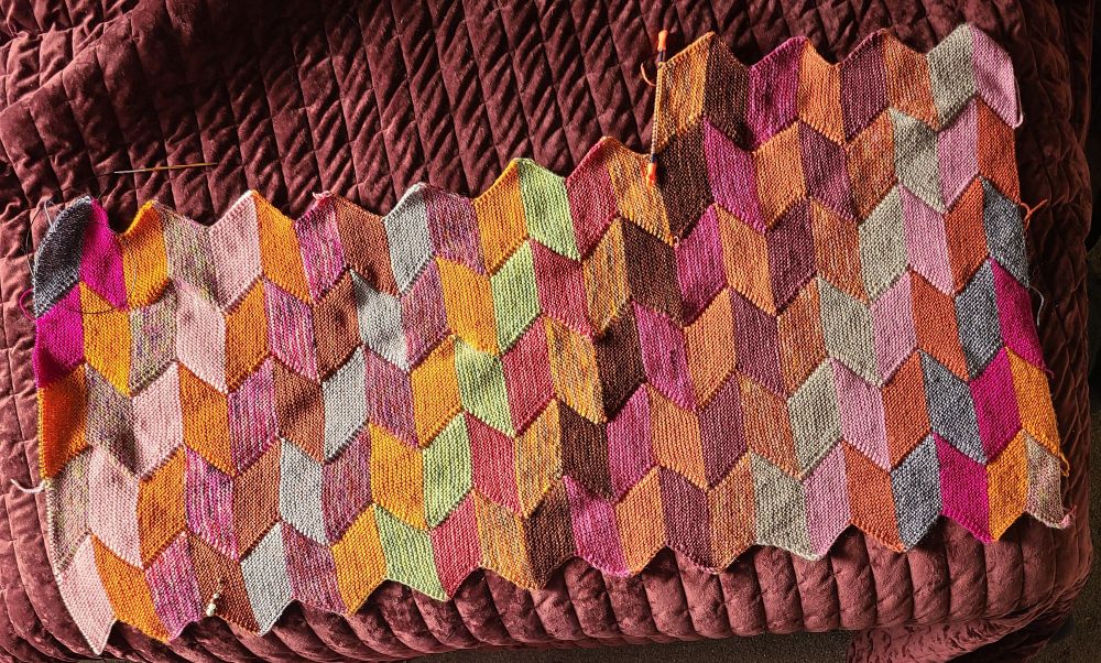 A blanket made of parallelograms. It has 22 colours to make a row and there will be 16 of these all together unless I run out of a colour before then. The colours rotate down as each new row is added with the first colour from the previous row becoming the final colour on the new row. Colours are all shades of orangey browns and pinks with a couple of grays, a green, and a blue to try and make it interesting. 20 out of the 22 colours are hand dyed.