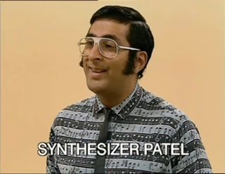 Picture of Synthesizer Patel, a 30-something Indian man with big glasses and sweet sideburns. He’s wearing a striped button up shirt with music notes and a very skinny black tie. He appeared in the TV series LOOK AROUND YOU.