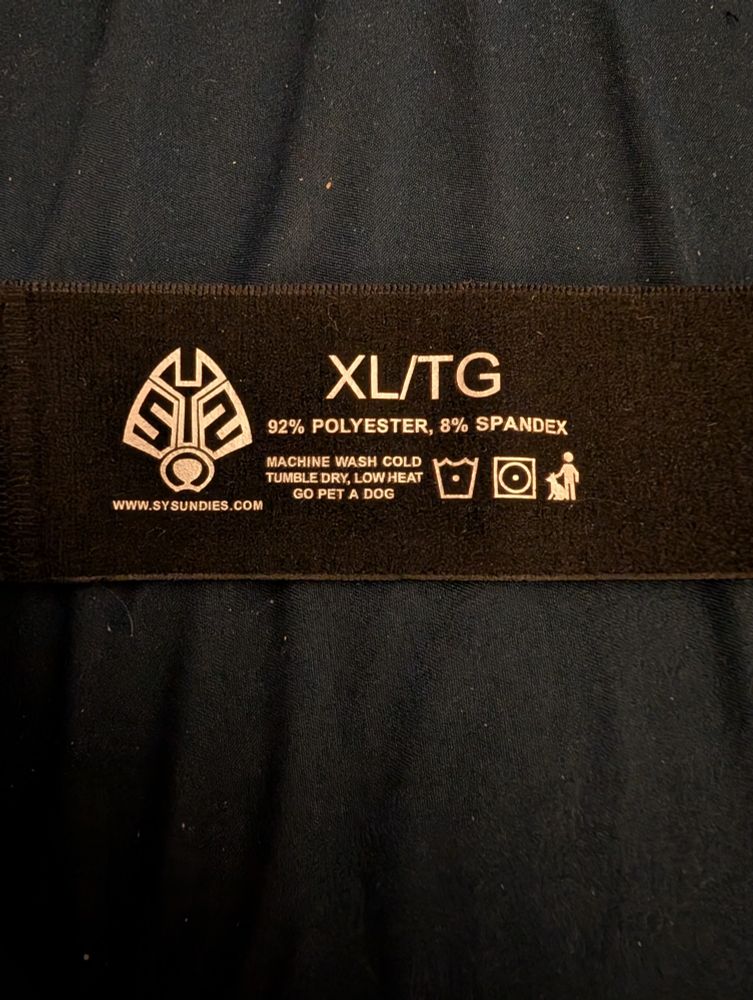 Tag on the back of the jock that reads, XL/TG, 92% Polyester, 8% Spandex.  Machine wash cold, tumble dry low heat, and to go pet a dog.  💜  The SYS Undies logo and website is also shown.