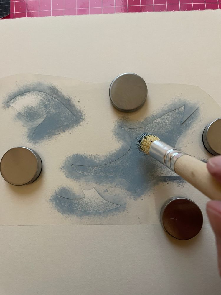Bristly brush applying blue-grey ink through plastic stencil onto paper. Small magnets hold the stencil in place 