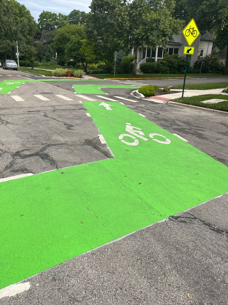 Depicts a bright green bicycle lane on a dark gray concrete street intersection with angled turns to follow how the street jogs. There’s a yellow caution traffic sign “watch for bicycles” on the right .