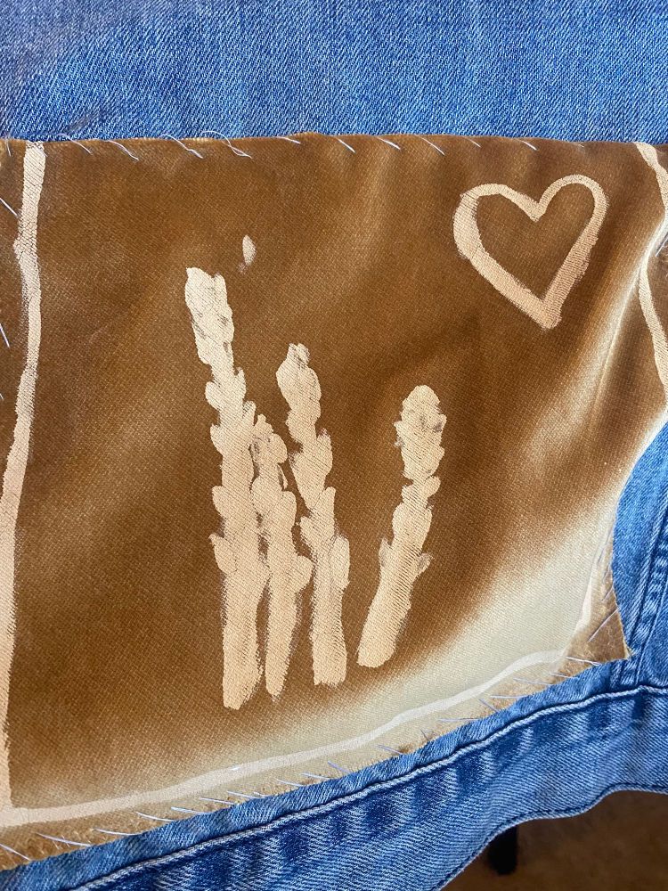 Fabric sewn on the back of a denim jacket. The patch is a gold color and velvety material. Painted in the center of the patch are four stalks of asparagus. A heart is painted in the upper right corner. There is a line on three of the edges of the patch, making a border. The painted elements are cream. The thread securing the patch to the jacket is light gray.
