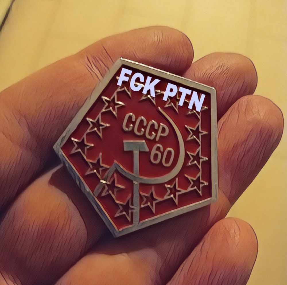 In einer Hand liegt ein fünfeckiger roter Metall-Pinn, goldfarben umrandet, in gold steht CCCP 60 geschrieben, darunter Hammer und Sichel. Der Rand des Pinns ist mit fünfeckigen Sternen verziert. Offensichtlich auf das Emblem hinzugefügt wurde die Überschrift FCK PTN.