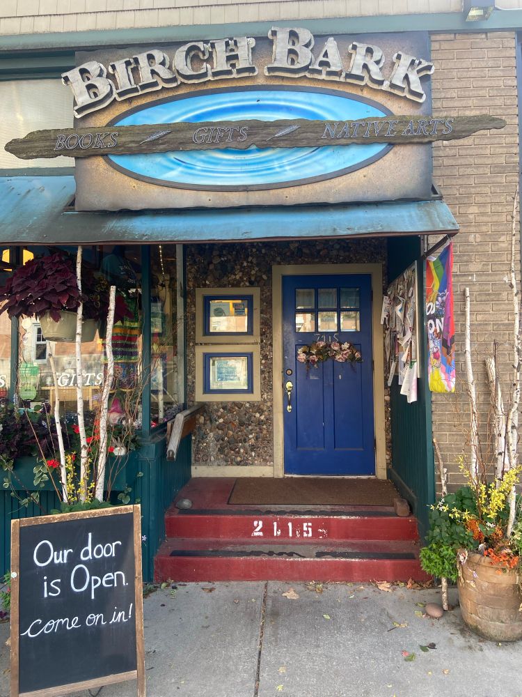 Outside View of Birchbark Books storefront. Blue door and awning visible along with chalkboard on sidewalk reading “Our door is open.”