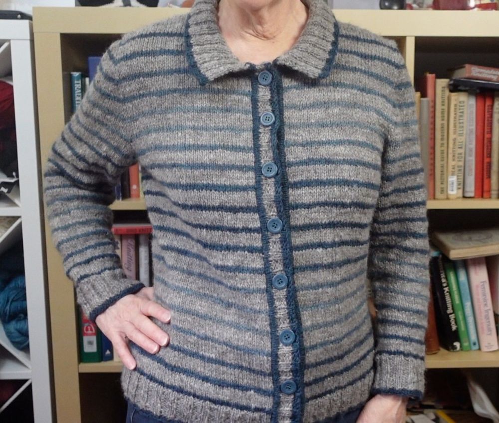 A woman is posed with one hand on hip in front of a bookcase full of knitting books. She is wearing a gray cardigan with a polo collar edged in dark teal yarn. The sweater has 2-row stripes of teal alternating with 5 rows of gray. The teal is in a gradient that shifts from darkest to lightest and back again several times from the shoulders to the bottom ribbing. There are five shades of the gradient. The button bands are edged with the dark teal at their base and at the cast off edge. There are 8 teal buttons down the front of the cardigan.