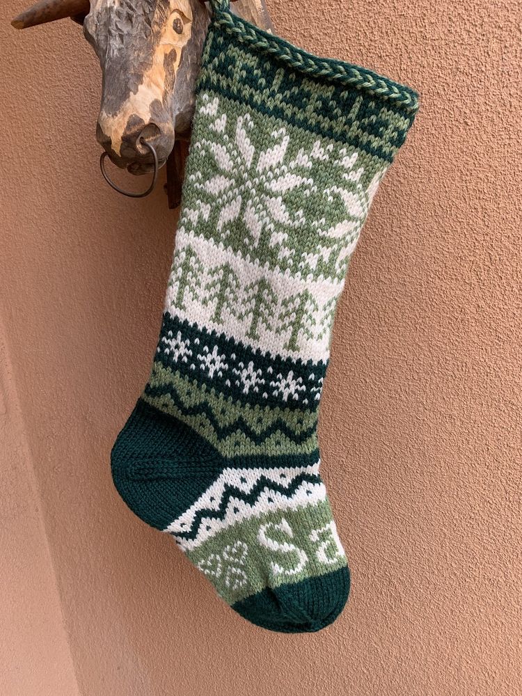 A Christmas stocking hangs against a rosy beige stucco wall. The stocking is knit in three colors: off white, sage green, and forest green. The top of the stocking has a Latvian braid knit with the two greens, and the stocking is knit in horizontal bands of stranded color work, using two colors for each band, with the width of the bands varying. The largest motif is a Scandinavian snowflake with sage green background and off white snowflake. Other bands have tree motifs, small snowflakes, and zig zag lines. The band on the foot is in sage green with "Sam" in white letters, flanked with three hearts. THe heel and toe are forest green.