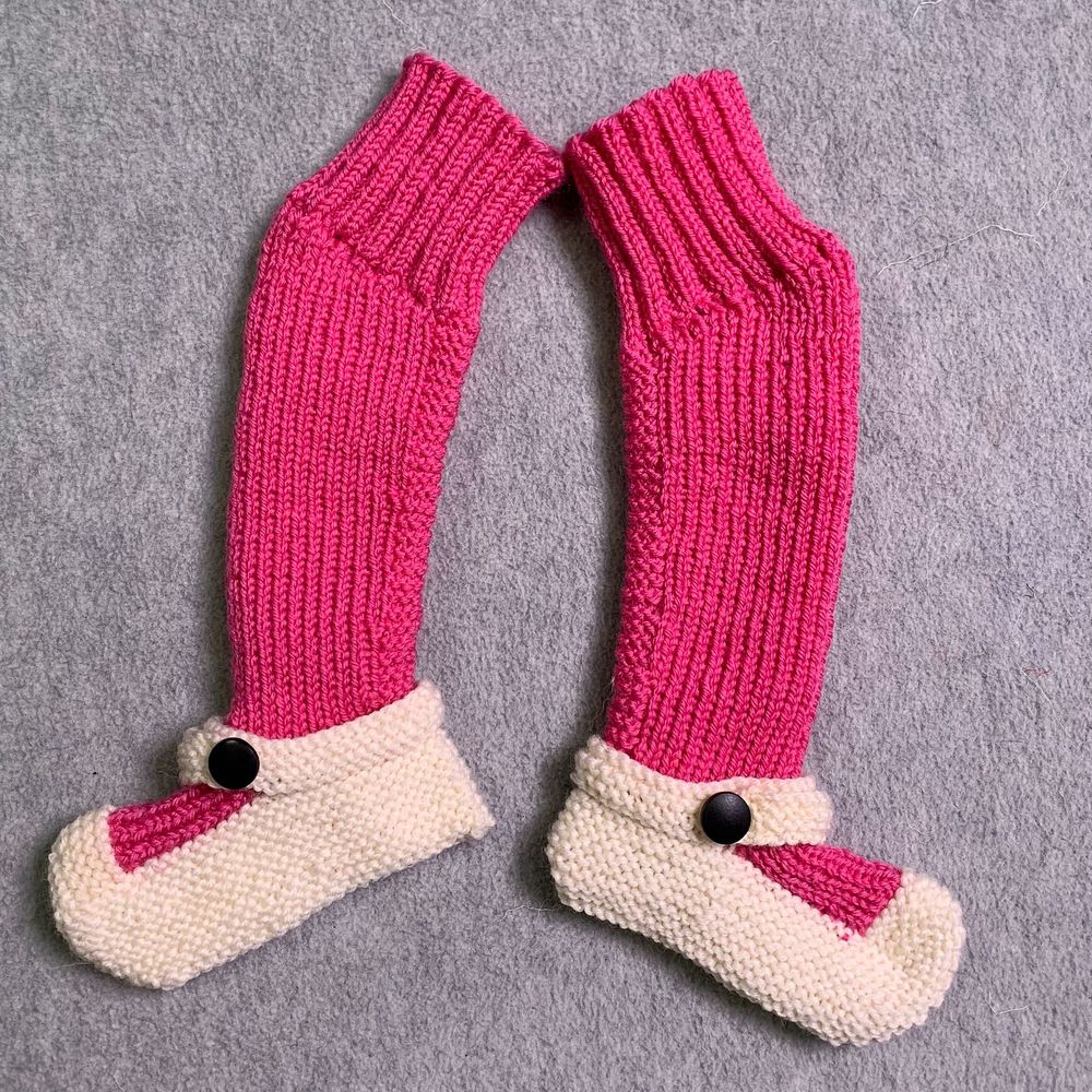 A pair of dark pink over-the-knee stockings lie on a gray background. The socks have short row shaping at the knee to create a natural bend, with k2p2 ribbing extending above the bend. The feet of the stockings are worked in garter stitch in a natural color to look like Mary Janes. A bit of the pink shows through between the toe and the  little ankle straps which have black buttons.