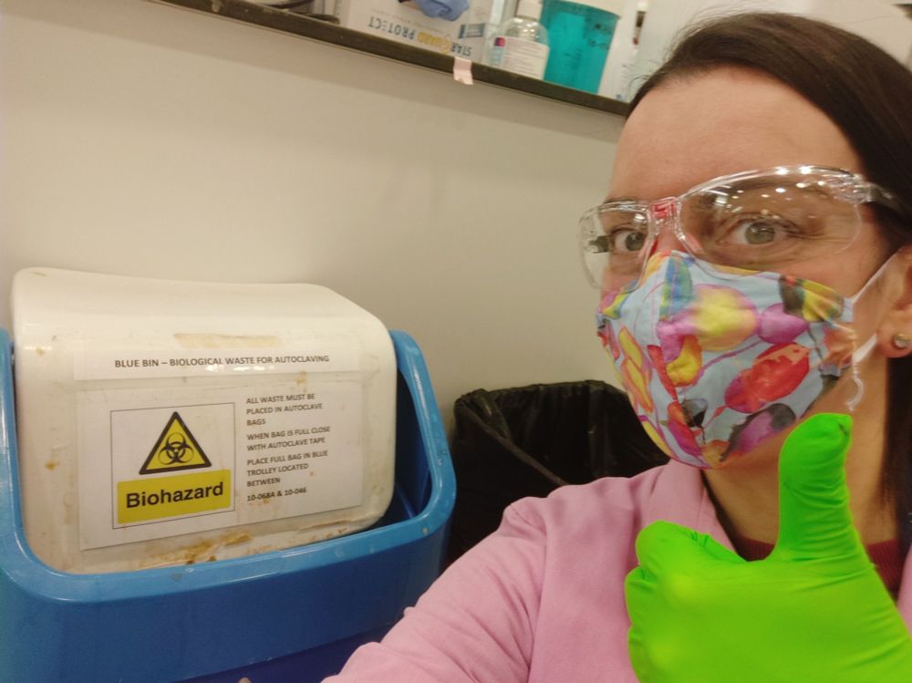 a woman wearing safety glasses, a mask, a pink lab coat and green lab gloves posing next to a trash bin labeled "biohazard". she's giving a thumbs up