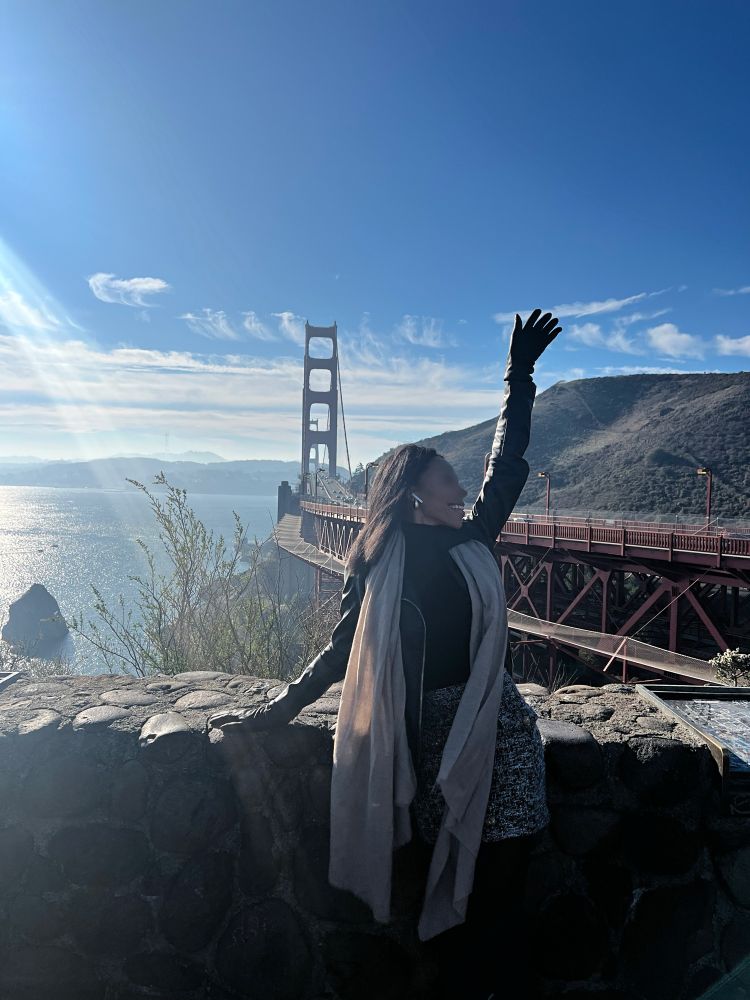 Nalani Belmont in SF posing in front of the iconic Golden Gate Bridge with an arm out and a happy expression on her face. She’s wearing a black leather jacket and a nude scarf around her neck, black panty hose and tweed black and white skirt and a black top. Very chic