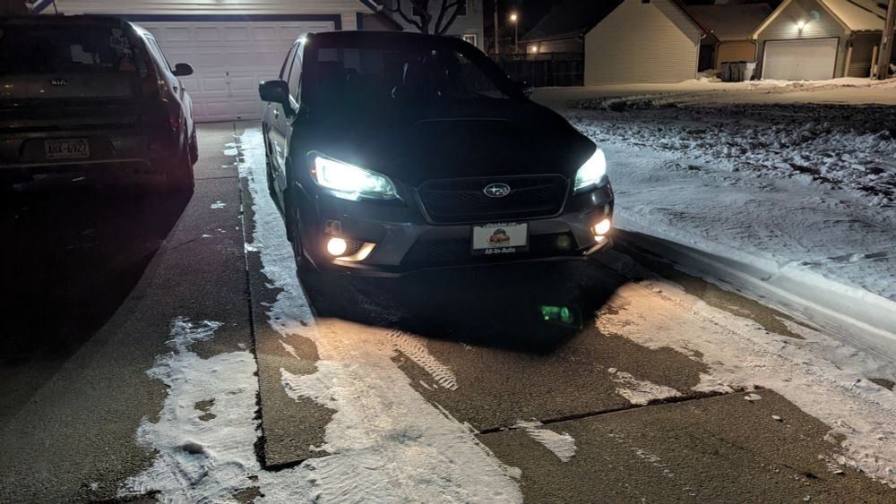 The front of a 2015 Subaru WRX in a driveway at night with the lights on.