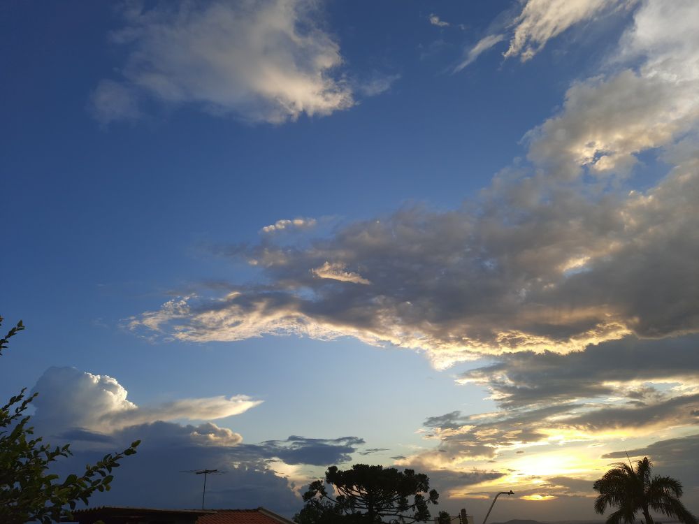 Uma foto do céu azul, com um por do sol no horizonte entre nuvens, algumas copas de árvores, um telhado e postes aparecendo no canto inferior em detalhe 